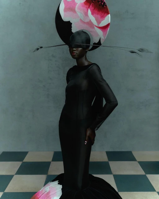 Fashionable African woman wearing a black gown and large floral headpiece poses in a muted-tone studio with checkerboard flooring.