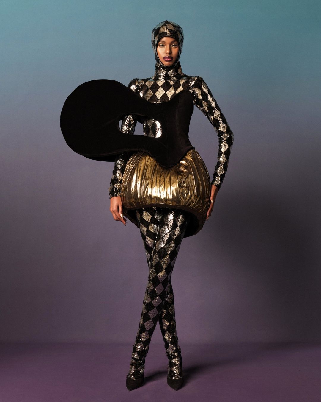 Full-length shot of a fashion model in a futuristic, harlequin-patterned costume against a blue and purple gradient background.