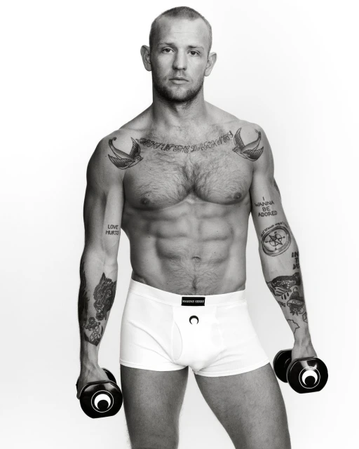 Black and white studio shot of a muscular man in underwear holding hand weights, showcasing tattoos on arms and chest against a white backdrop.