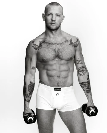 Black and white studio shot of a muscular man in underwear holding hand weights, showcasing tattoos on arms and chest against a white backdrop.