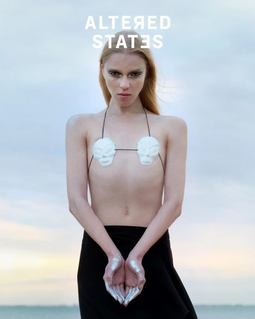 Female model in a conceptual fashion shot. She wears a black skirt and a mask bra, standing against a bright, dreamy sky background.