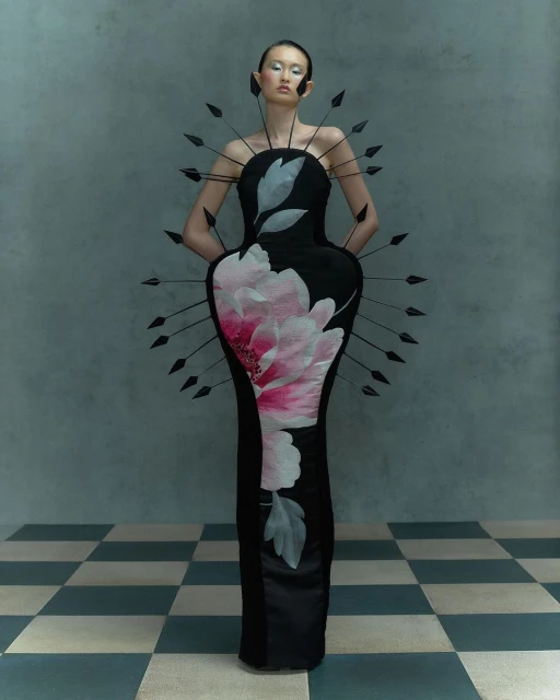 Elegant woman in a black floral gown posing in a studio with gray walls and checkered floor. Fashion shot with artistic composition.