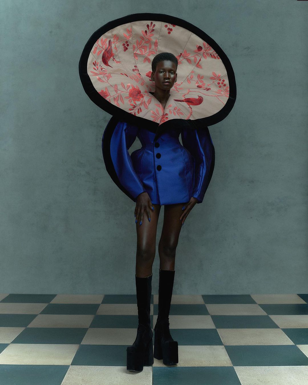 Woman in avant-garde fashion, featuring a blue dress with puffed sleeves, an oversized floral collar, black knee-high platform boots on a tiled floor.