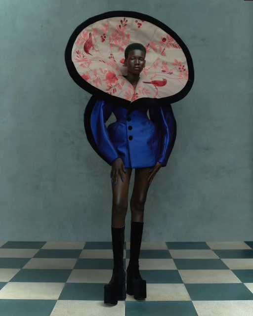 Woman in avant-garde fashion, featuring a blue dress with puffed sleeves, an oversized floral collar, black knee-high platform boots on a tiled floor.