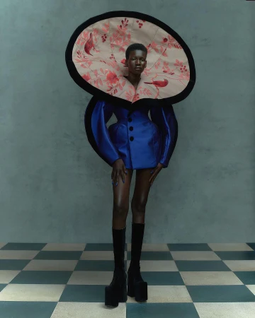 Woman in avant-garde fashion, featuring a blue dress with puffed sleeves, an oversized floral collar, black knee-high platform boots on a tiled floor.