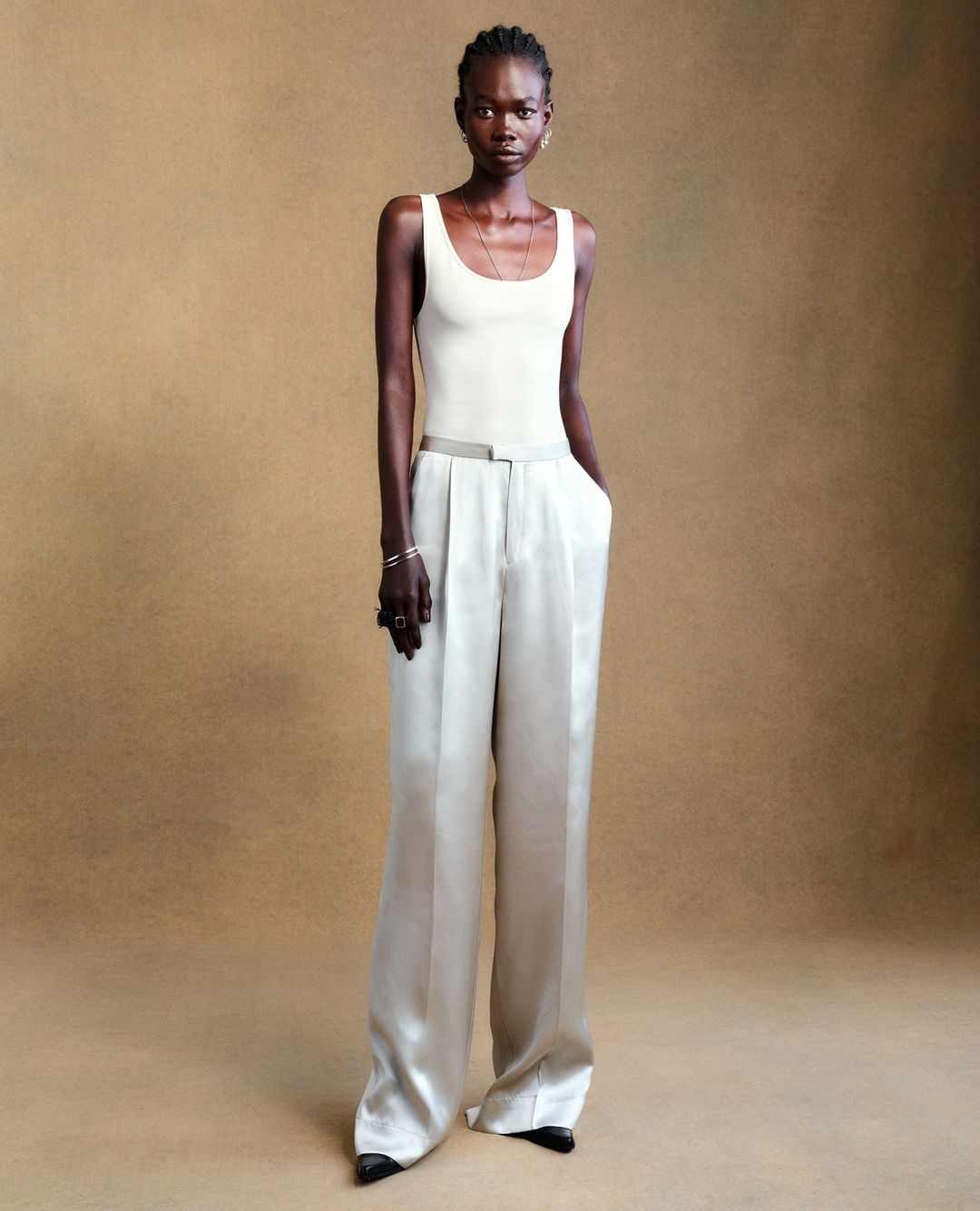 Indoor shot of a Black female model posing in a beige top and wide-leg pants against a plain beige backdrop in a fashion context.
