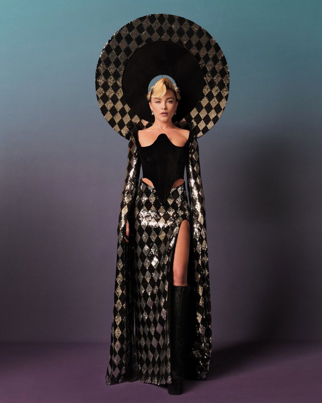 Full shot of a woman in a black and silver harlequin-patterned dress and matching hat, standing in front of a gradient background.