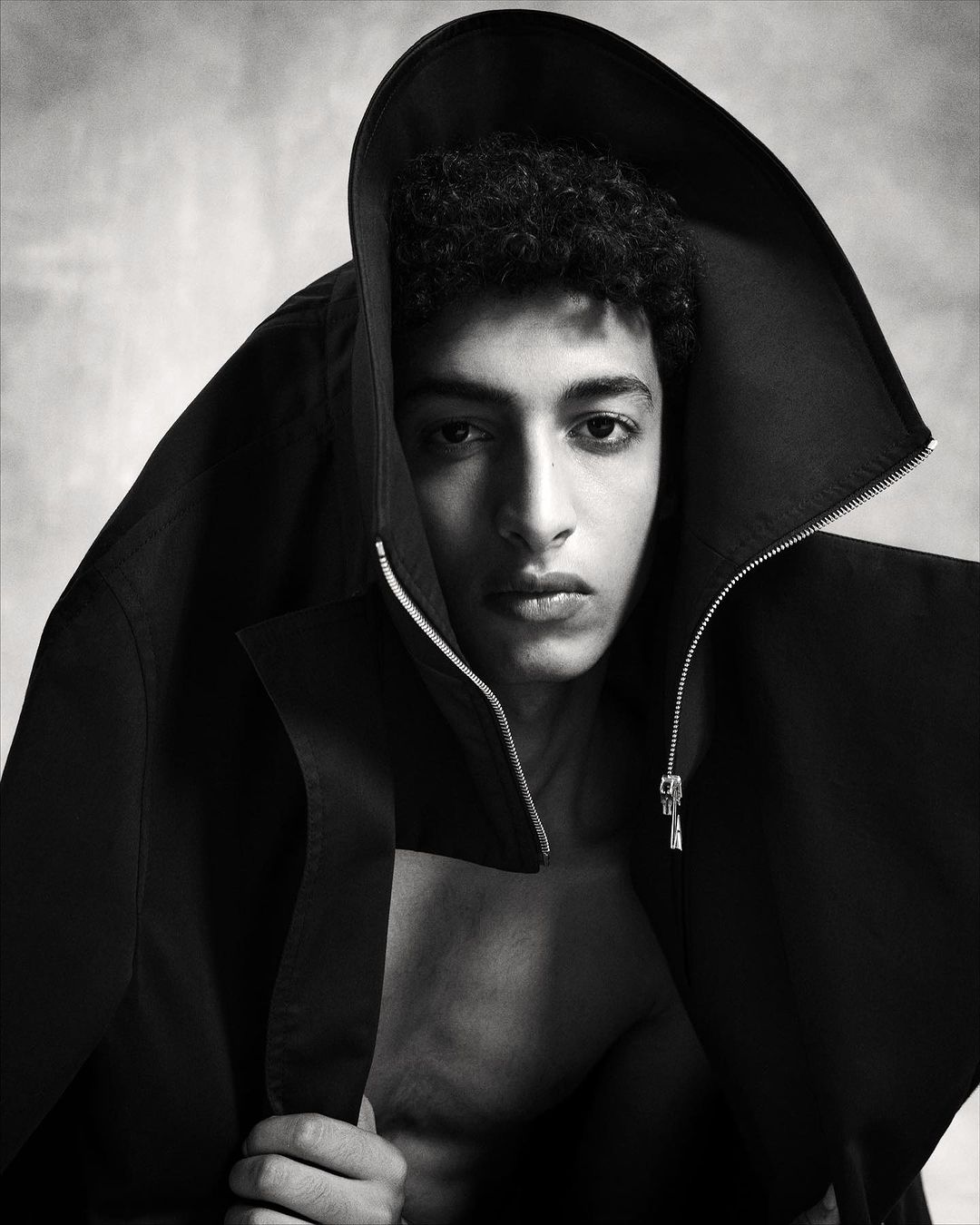 Portrait of a young man with dark curly hair wearing a hooded black jacket draped over his head, shot in monochrome by Marc Hibbert.