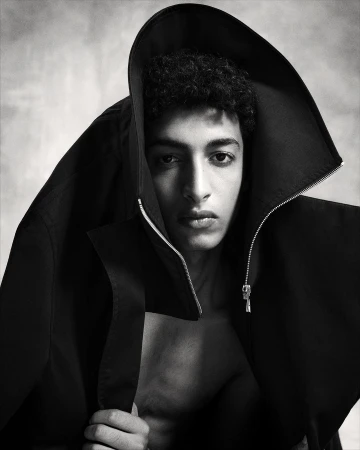 Portrait of a young man with dark curly hair wearing a hooded black jacket draped over his head, shot in monochrome by Marc Hibbert.