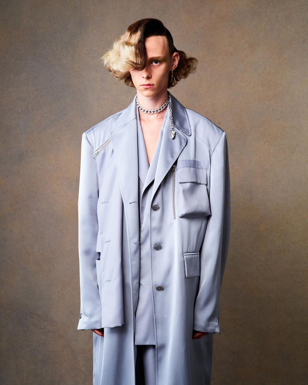 Portrait of a young male model with dyed hair wearing a light blue, oversized suit and silver necklace against a neutral background.
