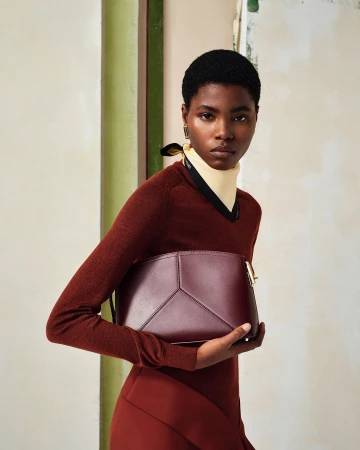 Model with short dark hair wearing a rust brown sweater and holding a dark burgundy leather handbag in a studio