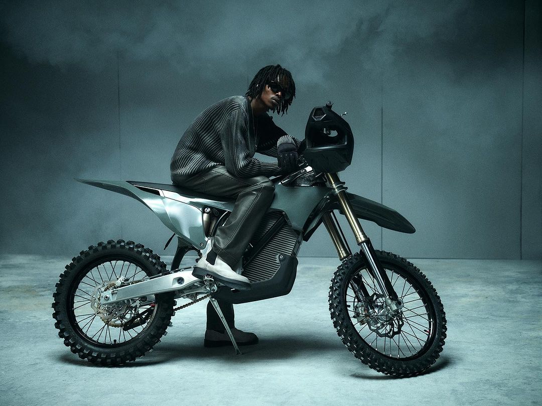 Man with dreadlocks rides silver electric dirt bike indoors wearing sunglasses and knit sweater in a grey studio with smoke effects.