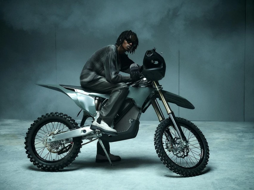 Man with dreadlocks rides silver electric dirt bike indoors wearing sunglasses and knit sweater in a grey studio with smoke effects.