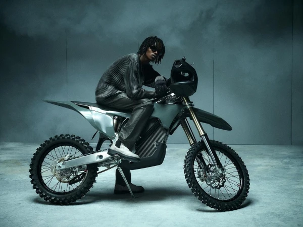 Man with dreadlocks rides silver electric dirt bike indoors wearing sunglasses and knit sweater in a grey studio with smoke effects.