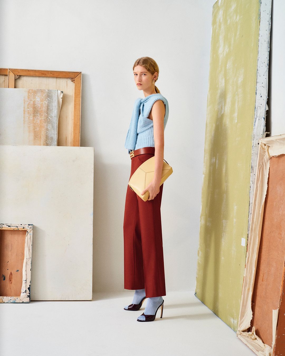 Fashion shot of a female model in red pants and a light blue top, holding a tan purse, posing in an indoor studio with painted canvas.