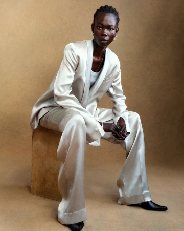 Portrait of a slender Black woman in a satin suit sitting on a wooden cube against a brown background in a fashion editorial style.