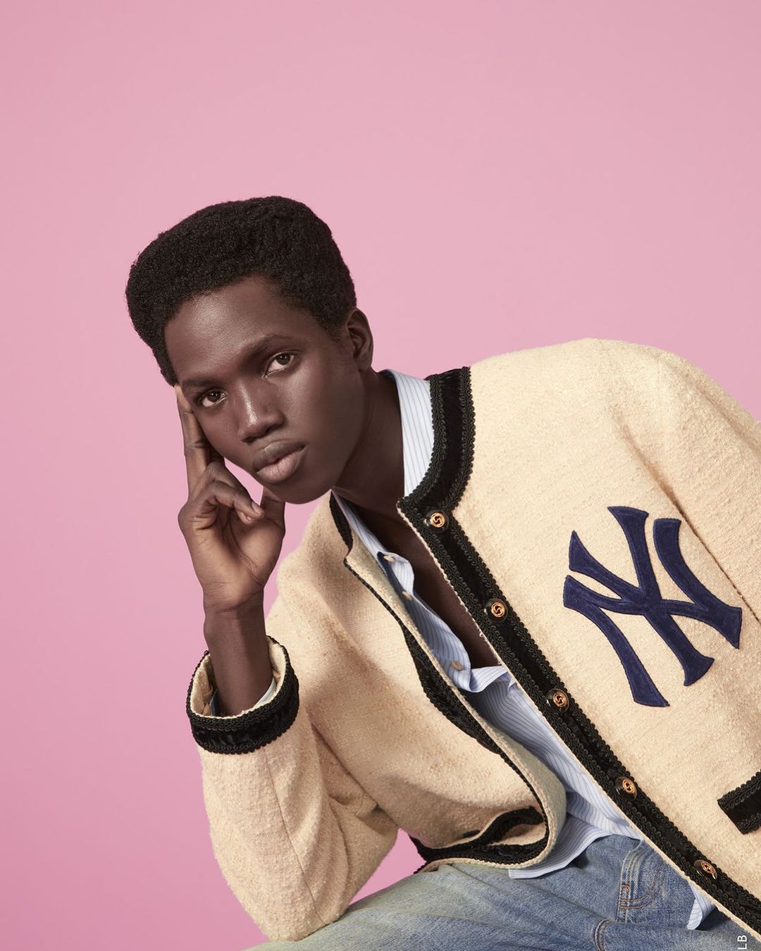 A portrait of an adult male model with dark hair, wearing a beige textured knit jacket with a navy blue baseball logo and blue jeans.