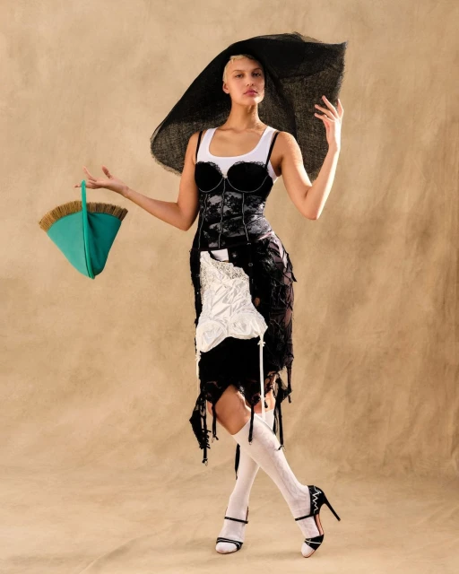 Fashion shot of a woman in a deconstructed dress with a large hat, holding a bucket-style bag, posing against a neutral background.