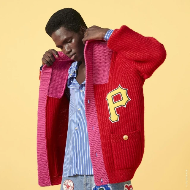 Young adult male putting on a red and pink knit varsity cardigan with an embroidered 'P' against a yellow background.
