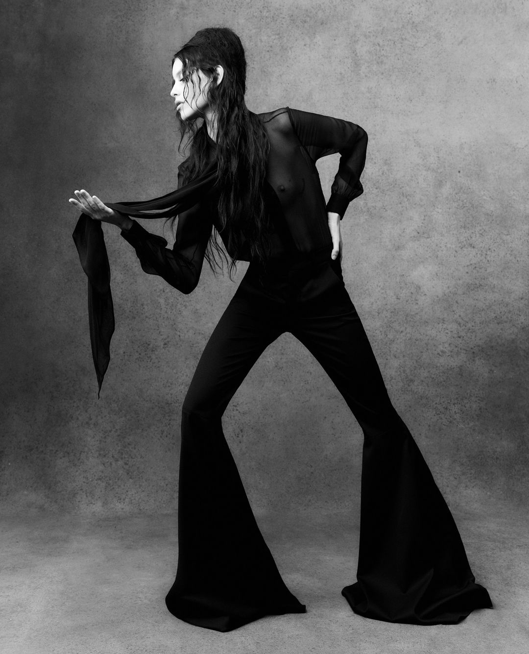 Black and white fashion portrait of a woman in a sheer top and flared pants, holding a scarf, against a textured grey backdrop.