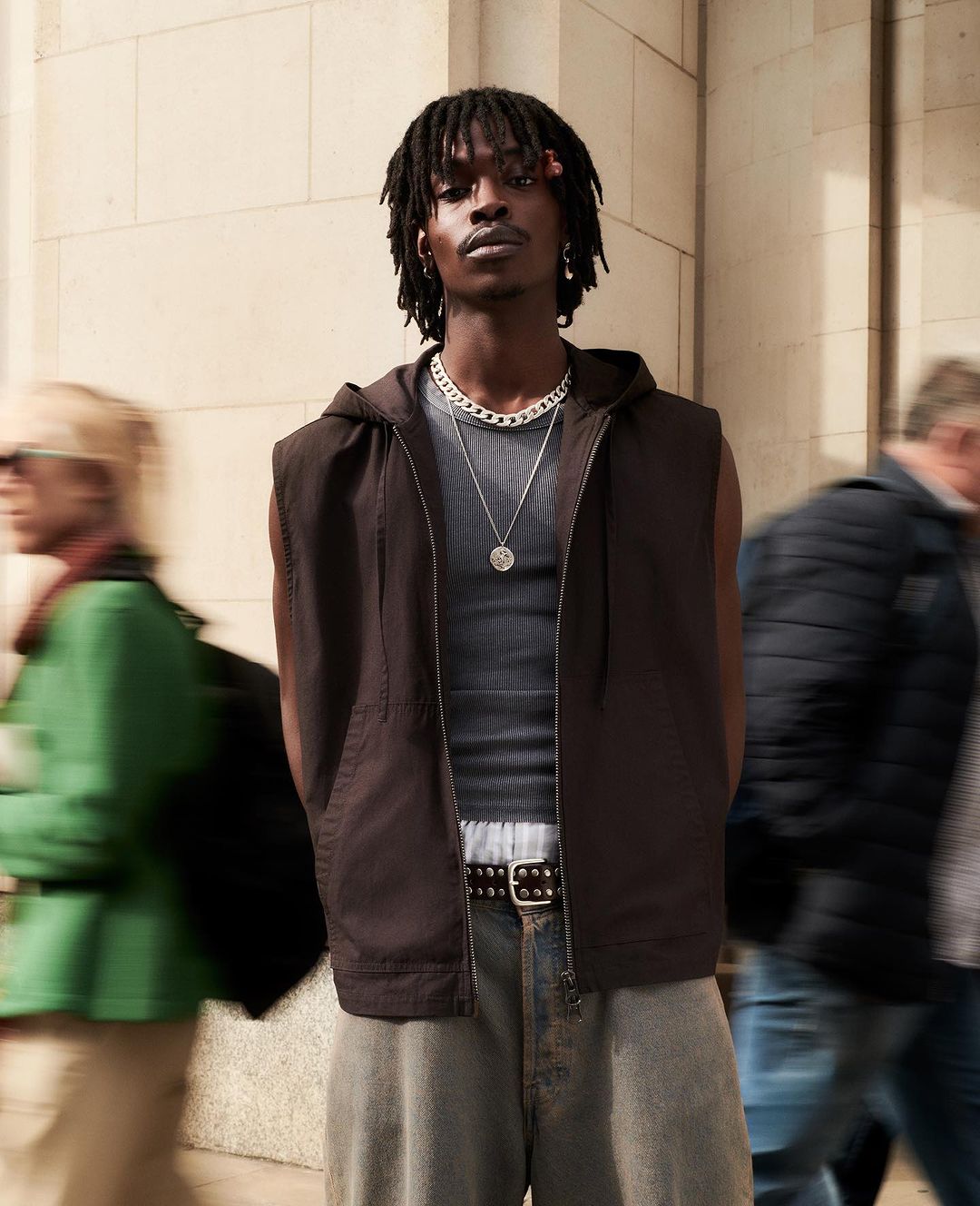 Man in a hooded vest and jeans stands outdoors with blurred people in the background. Fashion shot with natural light.