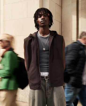 Man in a hooded vest and jeans stands outdoors with blurred people in the background. Fashion shot with natural light.