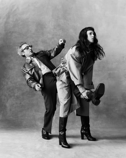 A man in a leather jacket prepares to kick another person wearing a tan trench coat and tall boots, shot against a neutral backdrop.