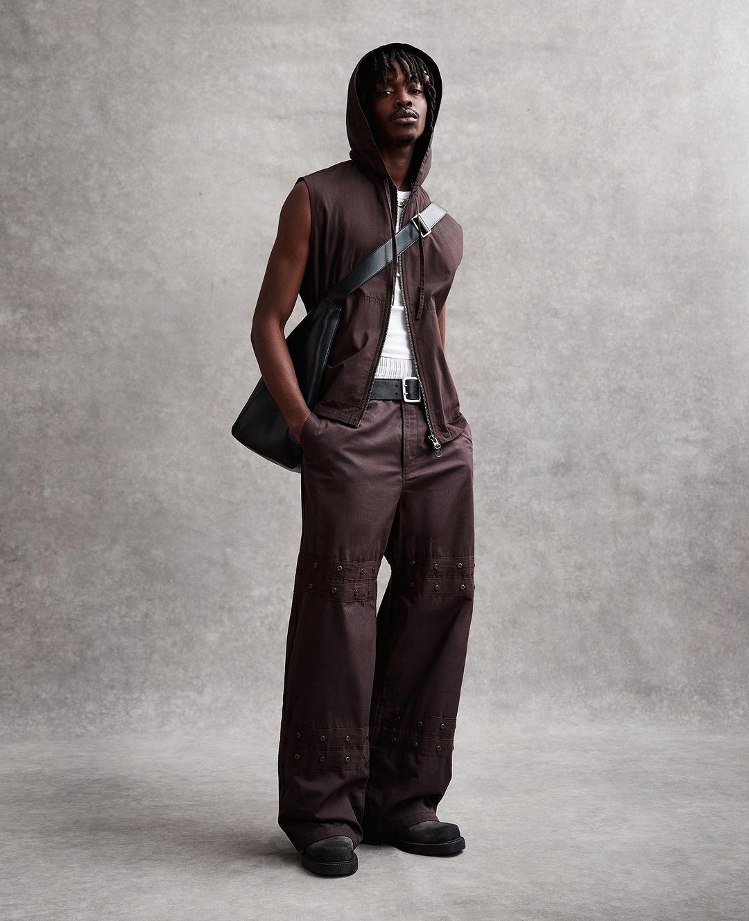 A young Black male model poses in a sleeveless hoodie, wide-leg brown pants, and black shoulder bag against a neutral gray backdrop.