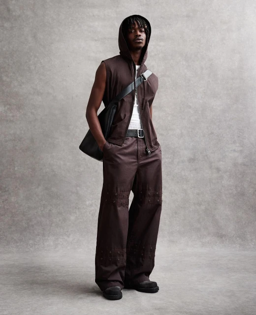 A young Black male model poses in a sleeveless hoodie, wide-leg brown pants, and black shoulder bag against a neutral gray backdrop.