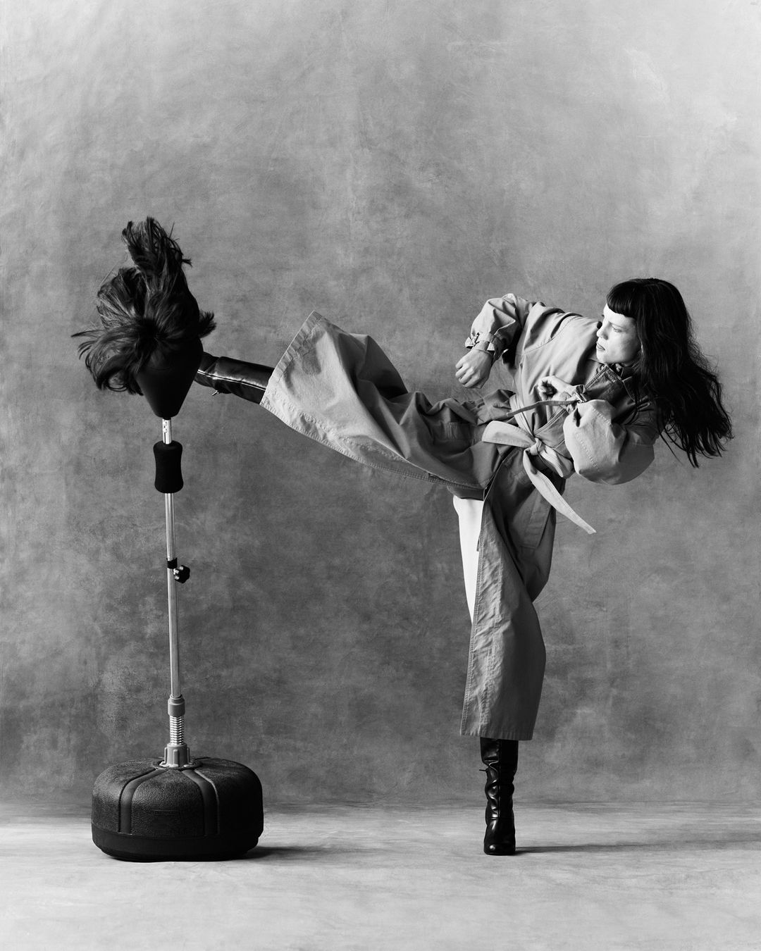 Monochrome portrait of a woman in a trench coat kicking a punching bag, showcasing fitness and fashion in a studio setting with soft lighting.