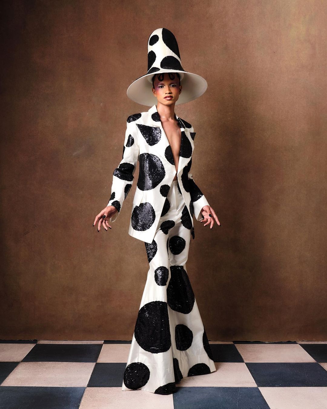 Fashionable person poses in a striking polka-dot suit and hat against a textured backdrop, standing on a checkered tile floor.