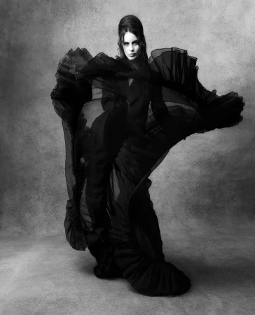 A woman in a full-length black gown poses dramatically with flowing ruffles against a textured grey background in a fashion photography style.