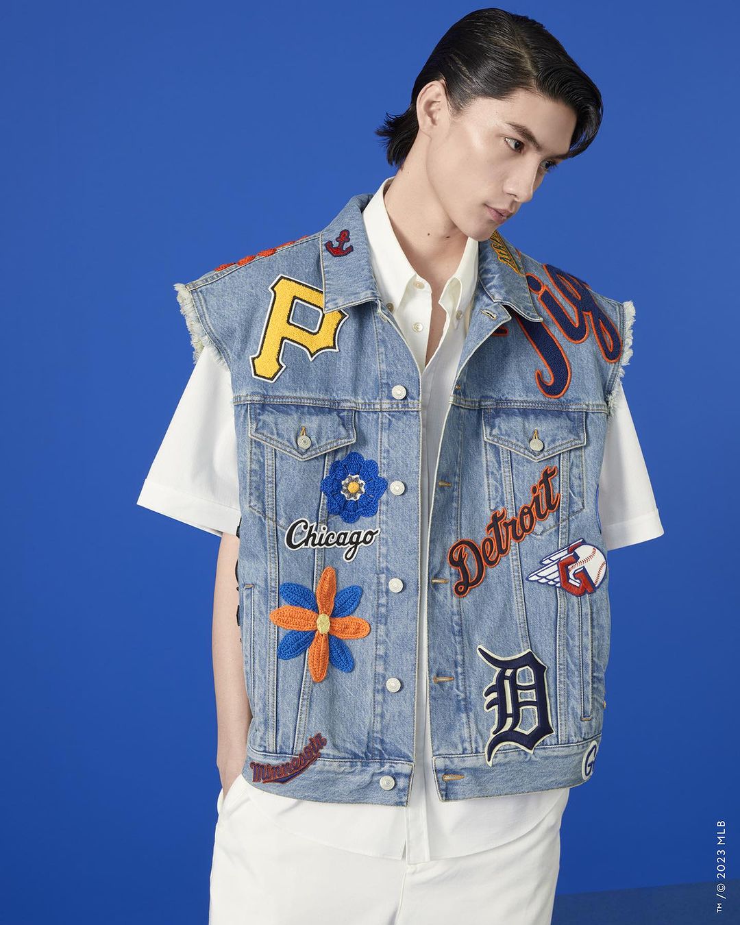 A person poses wearing a custom denim vest with patches and a white shirt against a bright blue backdrop in a medium studio shot.