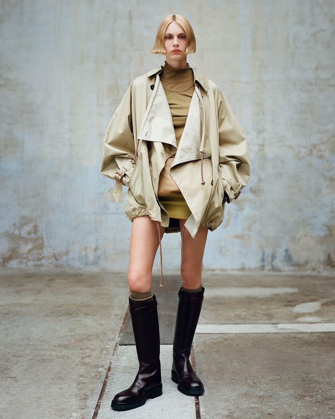 Full shot of a slender adult fashion model with fair hair wearing a trench coat and dark brown boots against a grey backdrop.