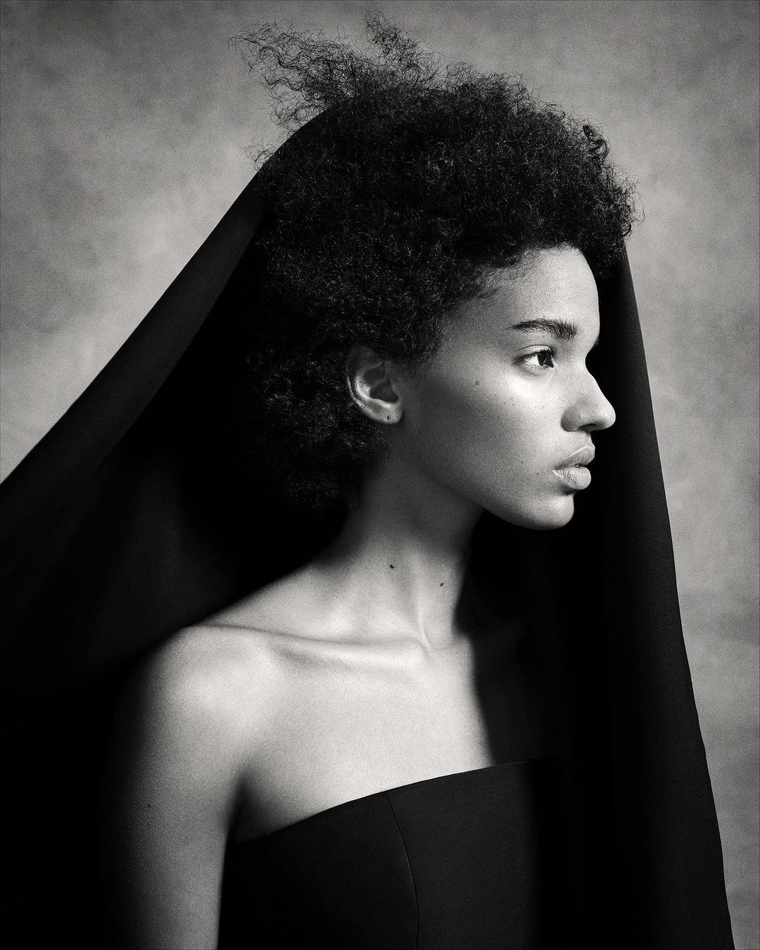 Monochrome portrait of a black woman in profile with a dark head covering and strapless black top against a mottled grey backdrop.