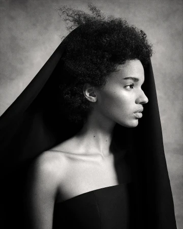 Monochrome portrait of a black woman in profile with a dark head covering and strapless black top against a mottled grey backdrop.