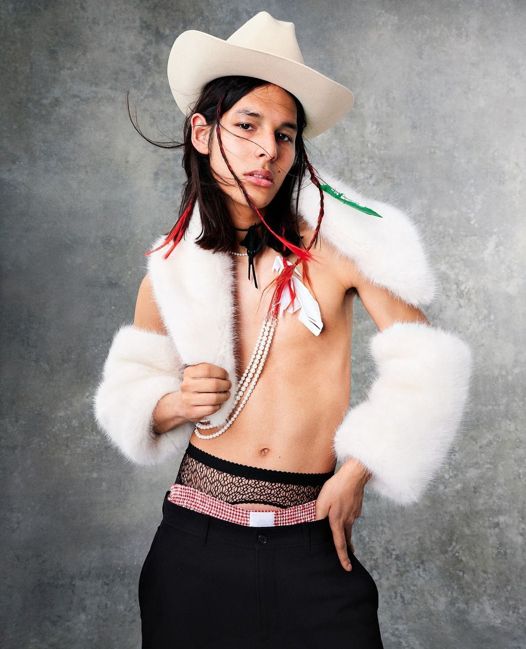 Man with long dark hair poses in cowboy hat, fur bolero, lacy underwear above black slacks, and pearl necklace against a gray backdrop.