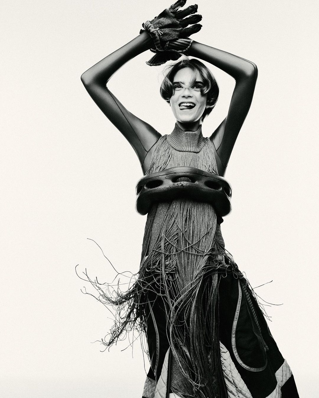 Monochrome studio portrait of a woman with short hair wearing an avant-garde fringe dress and gloves, striking a pose with arms raised.