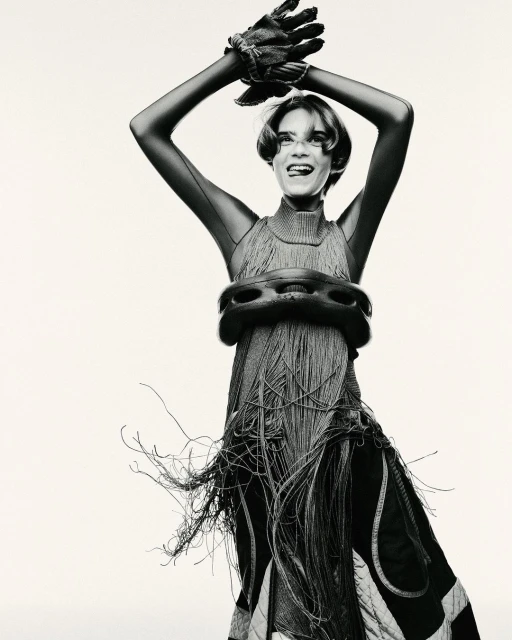 Monochrome studio portrait of a woman with short hair wearing an avant-garde fringe dress and gloves, striking a pose with arms raised.
