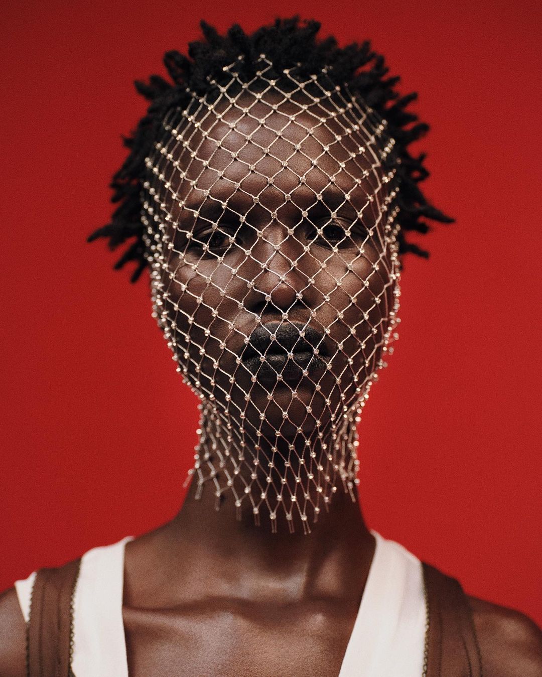Close-up portrait of a Black woman with dreadlocks wearing a white tank top and a beaded net over her face against a red background.