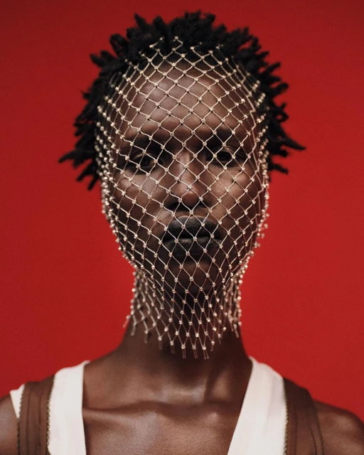 Close-up portrait of a Black woman with dreadlocks wearing a white tank top and a beaded net over her face against a red background.