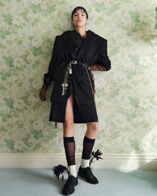A fashionable young adult woman poses indoors, wearing a black pinstripe suit with feathered accents, sheer socks, and embellished shoes against a floral wallpaper backdrop.