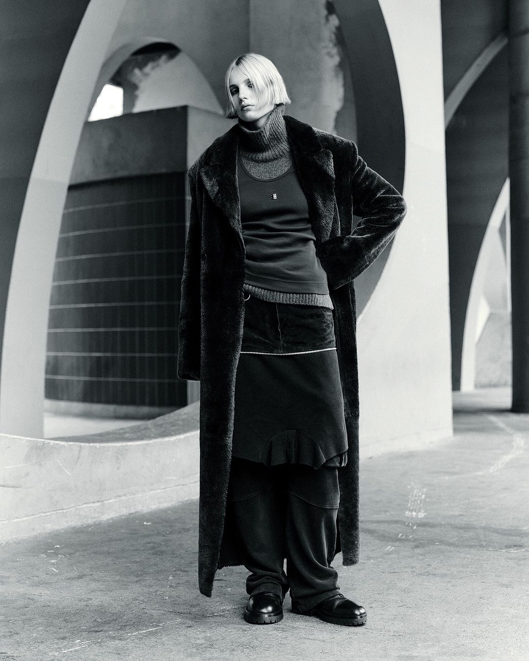 Monochrome full shot of a woman with blonde hair wearing a dark overcoat, turtleneck, skirt, and shoes, standing in a concrete structure.