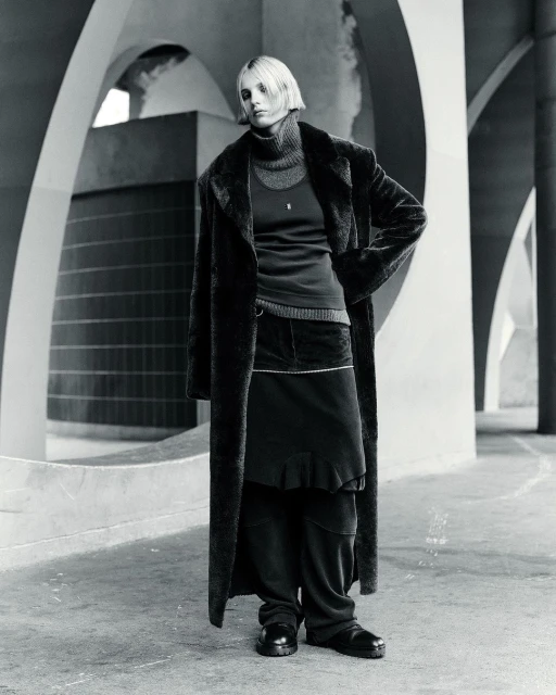 Monochrome full shot of a woman with blonde hair wearing a dark overcoat, turtleneck, skirt, and shoes, standing in a concrete structure.