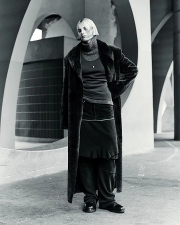 Monochrome full shot of a woman with blonde hair wearing a dark overcoat, turtleneck, skirt, and shoes, standing in a concrete structure.