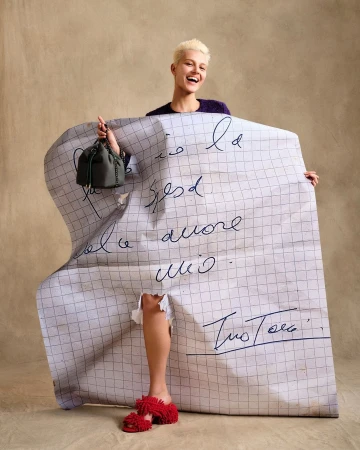 Model with blonde hair holding a large grid-patterned paper sheet with handwritten cursive text against a textured tan background.