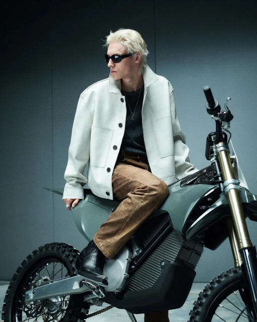 An adult male with blond hair and sunglasses sits on a gray off-road motorcycle in a photo studio wearing a light jacket and brown pants.