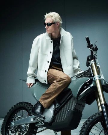 An adult male with blond hair and sunglasses sits on a gray off-road motorcycle in a photo studio wearing a light jacket and brown pants.