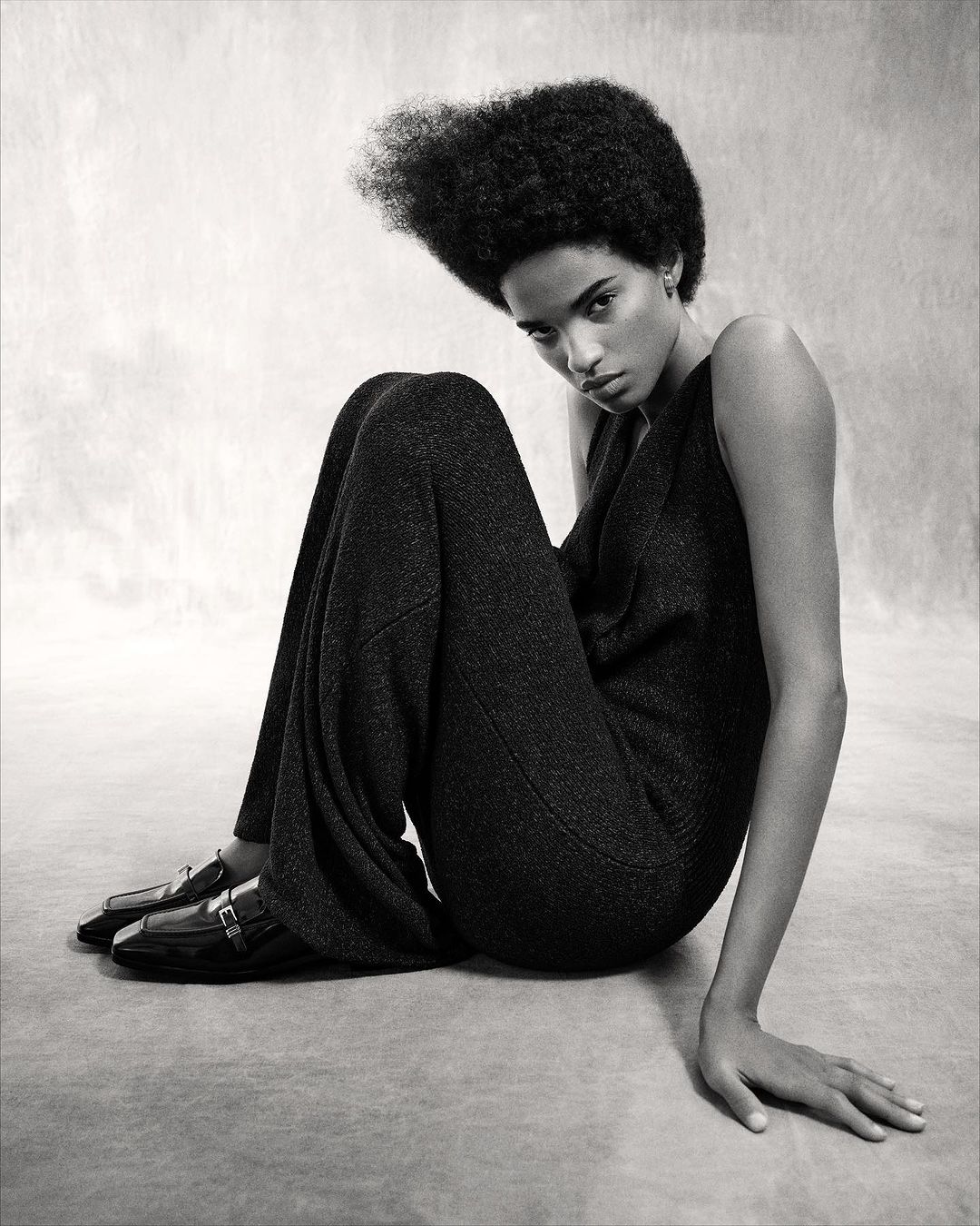 Elegant Black and White Studio Portrait of a Solo Adult Female Model with an Afro Wearing Dark Jumpsuit and Patent Leather Loafers.