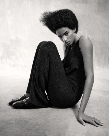 Elegant Black and White Studio Portrait of a Solo Adult Female Model with an Afro Wearing Dark Jumpsuit and Patent Leather Loafers.